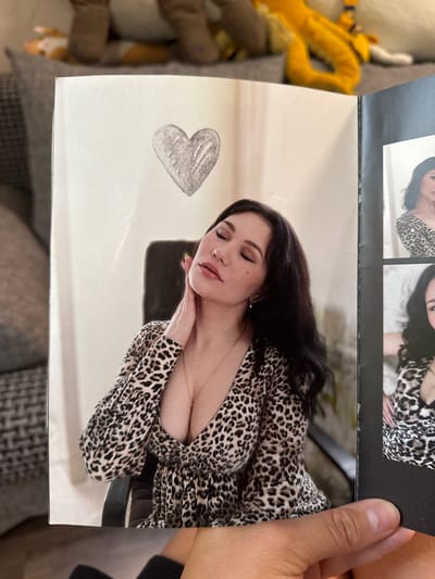 The image displays a hand holding a magazine open to a page featuring a photograph of a woman with dark, wavy hair and light skin, wearing a leopard-print dress. She has her eyes closed and her head tilted back, with one hand touching her neck. A drawn heart is visible above her head on the magazine page. To the right of this photograph, there are smaller inset photographs of the same woman in similar attire. The background of the main photograph appears to be an indoor setting with a white wall. The overall scene suggests a personal collection or display of photographs. There is no visible information or context that would allow for the identification of the city of Cheboksary, Russia.