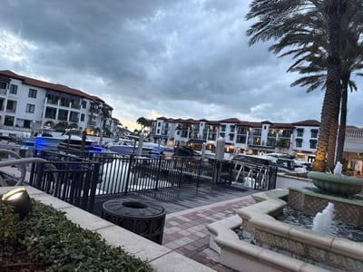 The image shows a waterfront scene in Naples, Florida, United States. Multiple boats are docked along a marina, some illuminated with blue lights. The marina is bordered by buildings with white facades and red-tiled roofs, typical of Mediterranean-inspired architecture. In the foreground, there is a stone fountain with water flowing, along with some greenery and a black trash receptacle. Palm trees are visible, adding to the tropical feel. The sky is overcast, indicating either dusk or an approaching storm. The setting appears to be a high-end residential or commercial area with waterfront access.