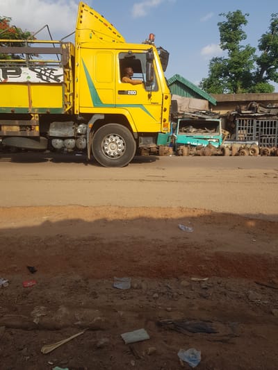 A large, bright yellow truck, identified by the number "260" on its side, is parked on a dirt road in front of what appears to be a vehicle repair or salvage yard. The truck's cab is occupied by a man wearing a light brown shirt. The yard behind the truck contains various vehicle parts, including axles and the chassis of a green truck. The overall setting suggests an outdoor scene in Kufena, Nigeria, possibly a commercial or industrial area. The road surface is rough and unpaved, with scattered debris and dirt. The sky is partly cloudy with a clear blue sky visible.