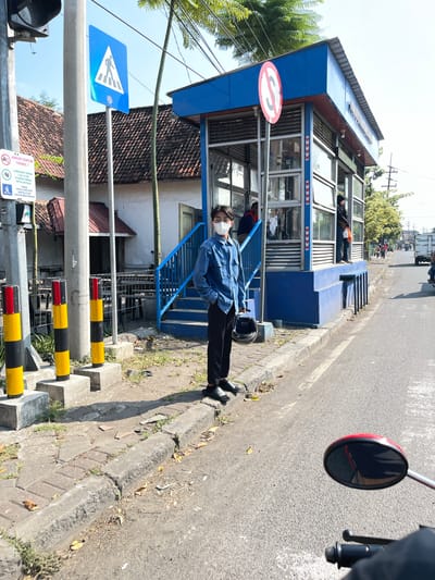 The image depicts a street scene in Kludan, Indonesia, likely near a bus stop. A young man wearing a blue denim shirt, black pants, a white mask, and black sandals stands on the sidewalk, facing the camera. He is holding a black helmet in his left hand. Behind him is a blue bus stop shelter with glass panels and blue stairs leading up to it. A pedestrian crossing sign is visible on a pole to the left of the man. Several yellow and black bollards are also present on the sidewalk. The street is paved, with a curb separating it from the sidewalk. In the background, other people are visible near the bus stop, and traffic, including a white truck and a motorcycle, can be seen on the road. The environment is urban, with buildings and trees visible. The sky is clear and blue.