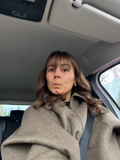 A female individual is seated inside a vehicle, located in the city of Mougins, France. She is positioned centrally, looking directly at the camera with her lips pursed forward. She has brown hair with bangs and wears a textured, light brown or taupe-colored coat featuring a silver zipper, with a black garment visible underneath. A black seatbelt crosses her right shoulder and chest. A gold-colored hoop earring is visible on her right ear. Above her, the vehicle's overhead console includes an "SOS" button and a green illuminated indicator light. A light-colored sun visor is partially visible on the upper right side of the frame. The exterior through the vehicle's windows shows ambient daylight, with visible moisture, consistent with rain, on the glass to the right.