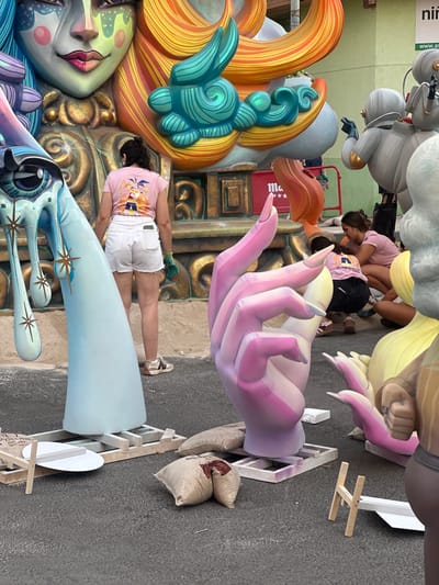 In Alicante, Spain, at the scene of the Fallas festival, large, colorful figures dominate the foreground and background. In the center, a woman wearing a pink t-shirt and white shorts stands facing away from the camera, looking at a massive sculpture of a face with vibrant blue and yellow hair, green and yellow makeup, and pink lips. To her left, a giant, pale blue, tear-shaped sculpture with golden star accents stands tall. To the right, a large, pastel pink and purple hand sculpture is prominent, with smaller figures and pieces of art scattered around. Several people are visible in the background, including a woman in a pink shirt and dark shorts kneeling and another person leaning towards her. The ground is paved, and there are sandbags and wooden supports placed around the base of the sculptures. A red barrier with a white logo featuring three stars is also visible. The overall scene depicts an outdoor street event with monumental, artistic sculptures characteristic of the Fallas celebrations.