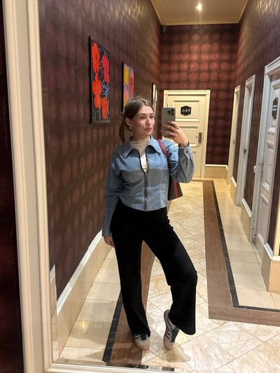 A young Caucasian woman with blonde hair pulled back in a ponytail is taking a mirror selfie in a hallway. She is wearing a light blue denim jacket zipped up over a white turtleneck, and loose black pants. She has on sneakers, and is carrying a dark-colored bag with a red strap over her right shoulder. Her left hand is holding up a smartphone to take the picture. The hallway has dark red, textured wallpaper with a repeating pattern, and framed artwork on the wall. There are several doors lining the hallway, some of which are open. The floor is tiled with a beige and cream marble pattern. The scene appears to be inside a building, possibly a restaurant, hotel, or public restroom area. There is no indication of the location being in Shkotovo, Russia.
