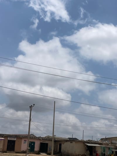 This media file depicts a street scene in Unguwar Saya, Nigeria, characterized by a partly cloudy sky. The image is oriented vertically, with utility lines and a power pole prominently featured in the foreground, stretching across the frame. These lines are suspended against a backdrop of blue sky interspersed with fluffy white clouds.
To the right of the utility lines, a row of buildings lines the street. These structures appear to be shops or residences, with some exhibiting signs of wear and tear. One building has a sign that reads "VICCALUX PAINT." The architecture suggests a typical urban or semi-urban environment in Nigeria. The lighting indicates it is daytime, likely late morning or early afternoon, given the brightness and the nature of the shadows. The weather appears fair, with the clouds suggesting potential for shade but not an imminent downpour. There are no people visible in the frame, focusing the viewer's attention on the built environment and infrastructure. The overall impression is one of everyday life in a Nigerian settlement, with a focus on the infrastructure supporting it.
