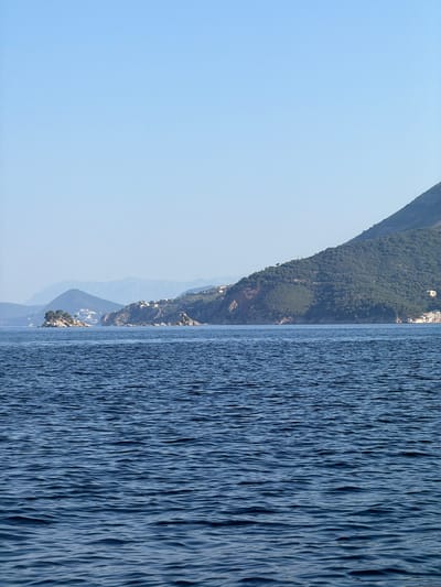 The image shows a panoramic view of the Adriatic Sea off the coast of Kaluđerac, Montenegro. The sea is a deep blue and has a slightly rippled surface. In the background, a chain of mountainous islands and the mainland are visible, covered with lush green vegetation and dotted with white buildings. The sky is a clear, pale blue. The overall scene conveys a sense of serene natural beauty and coastal habitation.