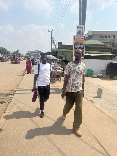The image depicts a daytime street scene in Ablekuma, Ghana, under a clear, hazy sky with strong sunlight casting long shadows towards the viewer's left. Two adult males are walking on a paved surface towards the right side of the frame.

The male in the foreground left, of dark complexion, wears a white short-sleeved t-shirt, black track pants with white stripes on the outer seams, and dark shoes. A dark backpack is visible on his shoulders, and he holds a burgundy item in his left hand. He looks generally forward.

The male in the foreground right, also of dark complexion, has a beard and mustache. He wears a light-colored short-sleeved shirt with a multi-colored bird and floral pattern, dark olive green pants, and light brown shoes. He holds a white smartphone in his left hand and appears to be looking towards the left, at the other man.

To their right, a utility pole stands prominently, featuring a multi-panel sign. The top panel reads "BIG CUCUMBER" with a monochrome image of a face. Below it, a larger sign advertises "DISCOVERY REMEDIAL CLASSES" with bullet points detailing "WASSCE MAY / JUNE," "NOV / DEC," "ONE YEAR WASSCE," "SHS VACATION CLASSES," and "COMPUTER CLASSES," along with a phone number "0550342555."

Behind the utility pole and men, a light green building with an air conditioning unit is visible. Adjacent to it are market stalls with white umbrellas, displaying goods. Other individuals are present in the background, including a person in a red garment walking away on the left side of the road. Further down the road, several vehicles, including a red pickup truck, are visible in traffic. Debris is scattered on the left side of the paved surface. Overhead utility wires traverse the sky.