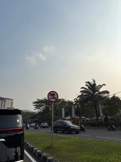 This is a street-level, outdoor photograph taken in Pakulonan Barat, Indonesia. The scene depicts traffic on a road during daylight hours, with a clear sky overhead. In the foreground, the rear of a black vehicle is visible on the left, with its taillights and a partial view of a sticker on the rear window. The road has multiple lanes, with several vehicles and motorcycles in motion. A traffic sign, a red circle with a diagonal bar across two stacked trucks, indicating "no trucks," is prominently displayed on the right side of the road.
Beyond the road, there is a grassy median with a palm tree and other trees, suggesting a green urban environment. In the background, there are buildings, including what appears to be a tall office building on the left, and further construction cranes can be seen in the distance. The lighting suggests it may be late afternoon, with a slight haze in the air. The overall impression is of a typical urban street scene in Indonesia.