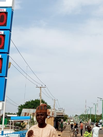 The image shows a street scene in Garko, Nigeria. On the left side of the frame, a tall blue structure displays red digital numbers indicating prices per liter, with "N/L" labels visible. In the foreground, a man wearing a light-colored traditional outfit and a patterned cap looks towards the camera. Behind him, a street stretches into the distance, lined with buildings, trees, and utility poles. Several people are visible walking and riding motorcycles on the street. A few vehicles, including a bus and cars, are also present. The sky is overcast.