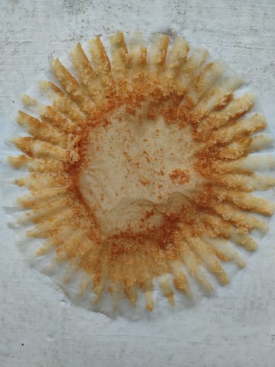 A single, circular, empty paper cupcake or muffin liner is centrally positioned on a light gray, textured surface in Rishtan, Uzbekistan. The translucent white liner retains its pleated structure, with its interior base and fluted sides extensively coated in light brown to reddish-orange granular residue, consistent with remnants of a baked good. The central portion of the liner shows less residue but exhibits a faint stain. No individuals or additional objects are visible within the frame. The background surface presents a uniform light gray hue with subtle textural variations.