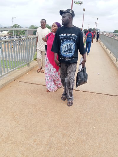 The image shows three people walking across a pedestrian bridge. The subject in the foreground is a man wearing a black t-shirt with a graphic design, dark washed jeans, and flip-flops. He is carrying a black bag in his right hand and a white envelope or thin bag in his left. To his left are a woman wearing a pink top and a patterned skirt, and a man in a beige shirt and pants, carrying a green bag. In the background, other people are visible on the bridge, as well as cars and buildings. The setting appears to be urban, with a cloudy sky, suggesting it might be daytime with overcast weather. The bridge has a metal railing on the left side, and the ground is paved with concrete. The overall impression is a casual scene of people commuting or traveling.
The t-shirt worn by the man in the foreground has the text "TENACITY" and "2002" visible on it. A woman in the background is wearing a blue uniform with "NSCDC" printed on the back.