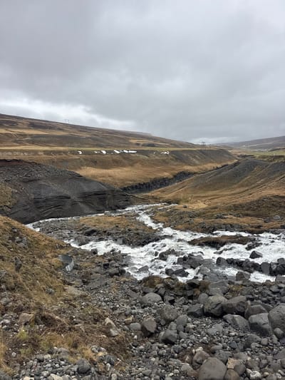 This image captures a rugged natural landscape under an overcast sky. A turbulent, white-foaming river or stream dominates the right side of the frame, flowing over and around numerous dark gray rocks of various sizes. The immediate banks and foreground are characterized by these scattered rocks and sparse, dry, brownish-yellow grass. In the lower right corner, a curved, metallic half-pipe, possibly a culvert or drainage pipe, is partially visible, embedded in the bank beside the water.

In the midground, undulating hills rise, covered predominantly in the same dry, straw-colored grass and dark, exposed rock. Patches of white snow are visible on some of these hillsides. Stretching across the midground, several utility poles or power lines are discernible as thin, dark structures against the muted landscape. The sky is entirely overcast, a uniform light gray, indicating a cloudy day with no direct sunlight. The overall scene suggests a cool or cold environment.

No people or animals are visible, and there is no explicit activity beyond the natural flow of the river. The lack of vibrant green vegetation and the presence of snow patches hint at a non-summer season or a harsh climate. There is no visible text in the image. The scene evokes a sense of wildness and desolation, consistent with a remote, natural setting.