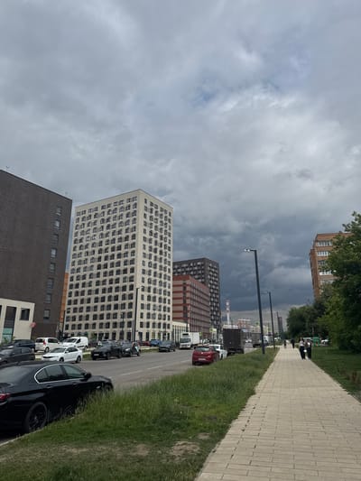The image depicts a street scene in Shcherbinka, Russia, under a cloudy sky. A paved pedestrian walkway runs along the right side of the frame, bordered by a strip of grass and then a street lined with parked cars. Several multi-story buildings, characteristic of urban residential areas, dominate the background. One prominent building is light-colored with a grid-like pattern of windows. Another, darker building, also with numerous windows, is visible in the foreground.

Several cars are parked along the street, including a black sedan in the foreground and various white and dark-colored vehicles further down. The sky is filled with heavy, dark clouds, suggesting overcast weather, possibly before or after rain. A few streetlights are visible along the walkway. In the distance, more buildings and what appear to be industrial structures or chimneys can be seen. The scene is quiet, with no overt activities or events taking place. The overall mood is subdued, reflecting the overcast weather. No legible text is visible in the image.