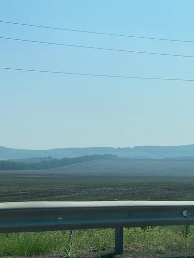The image depicts a rural landscape with rolling hills and fields under a clear, pale blue sky. In the foreground, a silver metal guardrail runs horizontally across the frame, with a patch of green grass and yellow wildflowers visible below it. The fields beyond the guardrail appear to be recently plowed or harvested, with dark soil. Further in the distance, a line of trees marks the base of the hills, which fade into a hazy blue in the background. There are three thin, dark horizontal lines stretching across the top of the sky, likely power lines. The scene is serene and devoid of any human presence or activity. The location is identified as Chulym, Russia.