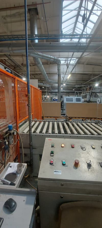 This is an indoor, medium-shot photograph taken from a low angle within an industrial facility, likely a manufacturing plant, in Kohtla-Järve linn, Estonia. The scene is dominated by machinery and infrastructure associated with a production line. In the foreground, a control panel with various buttons and indicator lights is visible. The panel has labels such as "RESET," "STOP," and "START," along with a sticker that reads "alldev.ce." To the left of the control panel, there's a grey metal surface with a black computer mouse and a small tray containing a marker and a circular object.
Behind the control panel, a conveyor belt system with multiple rollers stretches into the background. This conveyor appears to be transporting large, flat wooden panels. To the left of the conveyor, an orange metal safety fence is present, and beyond that, more industrial equipment and lighting are visible.
The ceiling of the facility features a skylight system that allows natural light to enter, illuminating the vast industrial space. Various pipes and ventilation ducts are suspended from the ceiling, indicating an active industrial process. The overall environment is utilitarian and functional, characteristic of a working factory floor. No people are visible in the frame. The lighting is a mix of natural light from the skylights and artificial lighting from overhead fixtures. The overall impression is one of a busy, operational industrial setting.