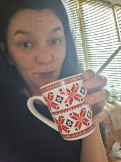 A woman with dark hair is holding a mug with a red and white snowflake pattern in her right hand. She is also holding a thin, cylindrical object, possibly a cigarette or a pen, between her index and middle fingers. The mug is positioned in front of her face, partially obscuring her mouth and chin. She is looking directly at the camera with her eyes open and appears to be indoors, near a window with venetian blinds. The background is slightly blurred, but suggests a domestic setting. There is no information to indicate this image is from Gotse Delchev, Bulgaria.