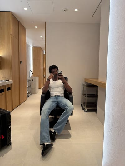 A man is sitting in a black chair in what appears to be a room interior. The man is light-skinned with short, curly black hair. He is wearing a white tank top and light blue denim jeans. He is holding a cell phone up, seemingly taking a picture of himself or recording a video. His legs are crossed, and he's wearing black shoes with white socks that are visible.
The background includes a clean, modern interior with light-colored walls and flooring. There are built-in wooden cabinets along one wall and some storage shelves visible near the ceiling. A black suitcase is located on the floor at the bottom of the frame. The location is undetermined as there are no signs to signify Fibracolor, Spain.