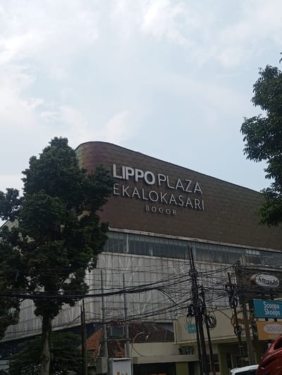 The image captures an exterior view of a large building under a bright, partly cloudy sky, with dense power lines and trees in the foreground.
The most prominent feature is the building, identified by the large white text on its upper facade: "LIPPO PLAZA EKALOKASARI BOGOR". The building has a curved or rounded corner on the left and a brown or dark tan exterior, possibly with a textured or patterned surface resembling small square tiles. Below the main sign, there are rows of windows, and the lower part of the building appears to have a lighter, possibly white or light grey, facade that looks somewhat unfinished or is covered with scaffolding-like material.
To the left, a large, leafy green tree partially obscures the building, extending from the bottom of the frame almost to the top left corner. Another tree is visible on the far right edge of the frame.
The lower half of the image is dominated by a complex web of black power lines and cables strung between multiple utility poles. These lines crisscross horizontally and vertically, creating a dense, tangled appearance. Below these cables, glimpses of other structures can be seen, including what appears to be rooftops of houses or smaller buildings with orange/brown tiles, and parts of what might be storefronts or ground-level businesses.
Several smaller signs are visible on the right side, partially hidden by the power lines. One white oval sign clearly shows the word "Amanda" in black script. Below it, a blue sign has text that appears to be "Scoop & Skoops" or similar, while a yellow or orange sign underneath that might say "DOH&GO". A red object, possibly part of a helmet or vehicle, is visible in the bottom right corner.
The sky is bright and a light blue, with scattered white clouds, suggesting it is daytime, likely morning or afternoon. The overall scene depicts an urban or semi-urban environment, characterized by the modern shopping plaza, dense utilities, and surrounding greenery.