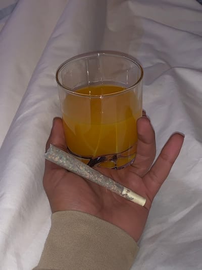 A hand holds a glass of orange juice and a rolled joint. The hand is partially visible, with a tan long-sleeve shirt covering the wrist. The juice is a vibrant orange in a clear glass with a visible handle. The joint is neatly rolled with a white filter tip and speckled green and brown filling. The background is a wrinkled white fabric, possibly a bedsheet.