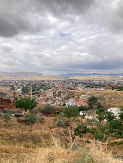 The image captures a panoramic view of Avanos İlçe Merkezi, Turkey, under a cloudy sky. In the foreground, dry, golden grass and scattered green trees cover a hillside dotted with rocks. A small outdoor seating area with a table and chairs is visible among the rocks.
The middle ground reveals a dense urban landscape with buildings featuring red-tiled roofs, interspersed with trees. Prominent structures include minarets of mosques and residential buildings. Some older buildings, appearing partially ruined or carved into the hillside, are also present.
In the background, a vast, arid plain stretches towards a range of flat-topped hills and mountains. The sky is overcast with dramatic, grey clouds. A Turkish flag is visible on a pole in the distance, indicating a public space. The overall scene depicts a town nestled within a dry, hilly landscape.