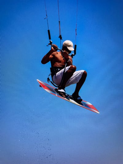 A man kiteboards in the air in front of a clear blue sky. He wears a white cap and white shorts. He holds onto the kite lines with both hands. His board is red, white, and blue. There are water droplets spraying to the right of the board. The location is likely Bacong, Philippines, based on the information provided.