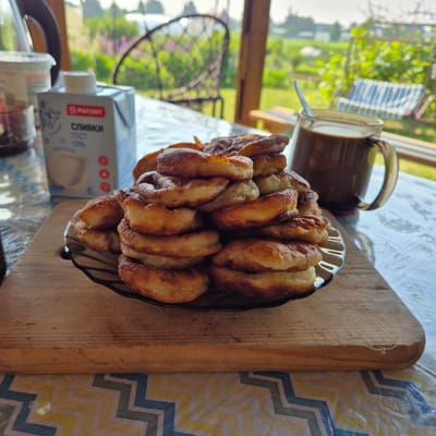 A close-up, eye-level shot captures a rustic breakfast scene on a wooden table outdoors, likely in a dacha or country house setting outside the city of Novo-Sroytka, Russia. In the foreground, a stack of golden-brown, fried pancakes sits on a dark, reflective plate, which is placed on a light-colored wooden cutting board. To the right of the pancakes, a clear glass mug filled with dark coffee, topped with a swirl of cream, rests on the table, a spoon extending from it. Behind the food and drink, a portion of a blue and white carton of "Magnit" brand cream is visible. The background offers a glimpse of a lush, green garden with scattered pink flowers and a partial view of a wrought-iron chair, suggesting a peaceful, rural environment. The overall impression is one of a simple, homemade breakfast enjoyed in a tranquil, outdoor setting.