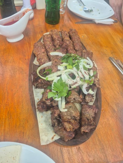The image captures a wooden platter featuring a generous assortment of grilled meats, possibly kebabs, garnished with raw onion rings and fresh parsley. The platter rests on a wooden table, likely outdoors. The setting is suggested to be Karatakla, Georgia. Elements visible around the platter include a white sauce boat, bottles, and a partly eaten plate.