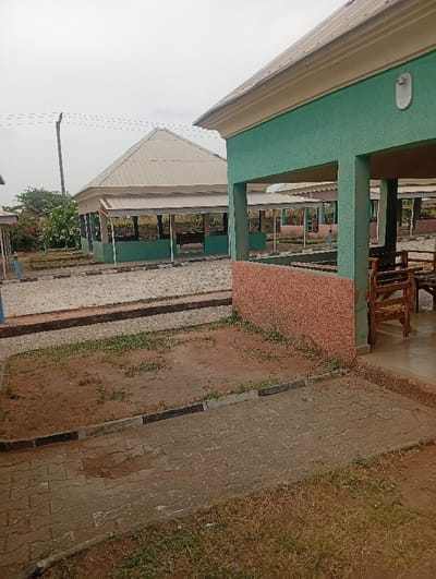 This image depicts an outdoor communal or recreational area in Kado, Nigeria, under an overcast sky. In the immediate foreground on the right, a structure with green-painted walls and a cream fascia features an open-air section supported by columns, revealing wooden benches and tables inside. The lower portion of this structure's wall is finished with a textured reddish-brown material, and a white outdoor light fixture is mounted on the green wall.
A paved pathway, constructed from interlocking gray pavers, extends from the foreground, bordered by curbs with alternating black and white paint. To the left of this pathway, an area of bare earth and sparse green vegetation is visible. In the midground, a prominent gazebo-like structure with a light-colored corrugated metal hip roof and green-painted base walls stands centrally, also appearing open-sided with interior seating. To the far left, a utility pole supports multiple overhead power lines, and dense green foliage with scattered pink flowers is present. Additional similar open-sided structures are visible further in the background. The ground surfaces exhibit a mix of worn paved sections, exposed soil, and sporadic plant growth. No individuals or active subjects are present in the scene.