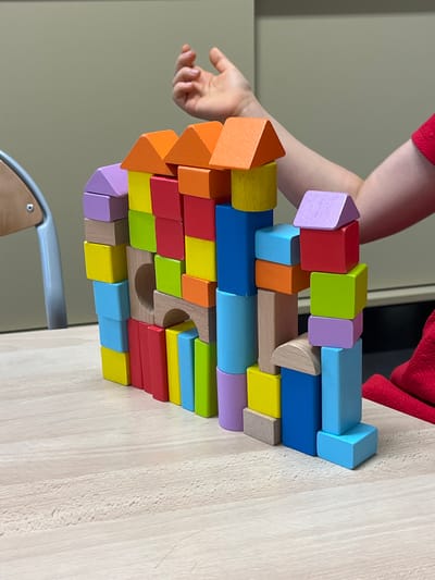 The image depicts a colorful tower of wooden blocks constructed on a light-colored wooden table. The blocks are of various shapes and sizes, including rectangular prisms, cubes, and triangular prisms, and are painted in vibrant colors such as red, orange, yellow, green, blue, and purple. A small, circular opening is visible in the center of the block structure. In the background, a person's arm and hand are visible, suggesting they are interacting with the blocks. The person is wearing a red garment. The environment appears to be indoors, with a neutral-colored wall in the background and a part of a chair visible on the left. The image does not contain any elements that would indicate a specific location in the city of La Fouillouse, France.