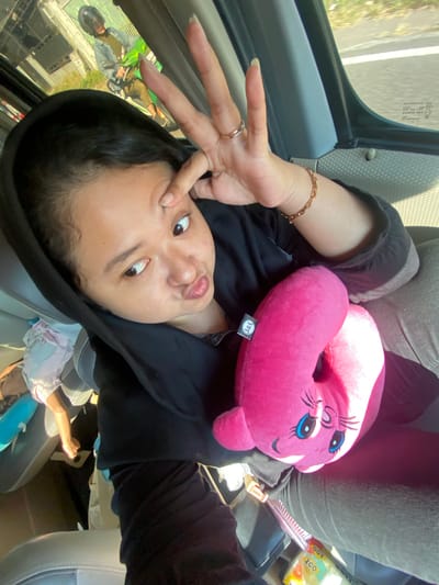 A woman, wearing a black hoodie, is sitting in a vehicle. She is looking at the camera with her mouth pursed and is holding up two fingers in a peace sign to her forehead. She is holding a pink stuffed animal in her lap. In the background, outside the vehicle, another person is riding a motorcycle. The scene appears to be a street in Pacing, Indonesia.