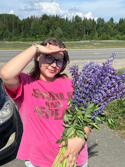 A young woman stands outdoors holding a large bouquet of purple lupine flowers. She is wearing a bright pink t-shirt with a graphic print and black-rimmed glasses. Her right hand is raised to shield her eyes from the sun, and she is looking towards the viewer with a slight smile. To her left, the front of a dark-colored car is visible. The background shows a paved road, a line of green trees, and a blue sky with scattered clouds. The image appears to have been taken on a sunny day in a rural or roadside area. The location is identified as Bolshaya Usa, Russia.