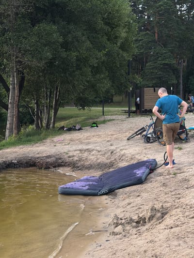 In Malakhovka, Russia, a man is seen on a sandy embankment near trees and a body of water. He is wearing a blue t-shirt and brown shorts, and appears to be using a foot pump connected to an inflatable mattress or sleeping pad. The mattress is dark purple with a subtle pattern. A bicycle is parked nearby. The scene suggests an outdoor recreational activity, possibly camping or preparing for an outdoor adventure by the water. The lighting indicates it is daytime, and the weather appears to be mild. In the background, there are more trees, a small wooden structure, and other people, suggesting a public area like a park or beach. Some personal belongings, including a backpack and a pink item, are scattered on the sand in the middle ground. The "Intex" logo is visible on the inflatable mattress.
