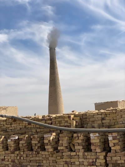 This image depicts an active brick kiln site in Nakhonay, Afghanistan, under a partially cloudy blue sky. A tall, light brown, tapering chimney dominates the central background, emitting a plume of grey smoke from its top. In the foreground, numerous light brown and reddish bricks are neatly stacked in organized columns, forming a substantial wall-like structure that extends across the lower half of the frame. A thick, dark pipe, possibly a water or drainage line, runs horizontally across the mid-section of these stacked bricks. To the left, behind the brick stacks, a low, flat-topped mud-colored structure is visible. A small, dark, indistinct figure is discernible near the base of the chimney, positioned behind the foreground brick stacks and to the right of the leftmost structure. To the right of the chimney, another mud-colored structure, possibly with a rudimentary roof, is partially visible above the brick stacks. No direct interactions or specific actions beyond smoke emission are discernible.