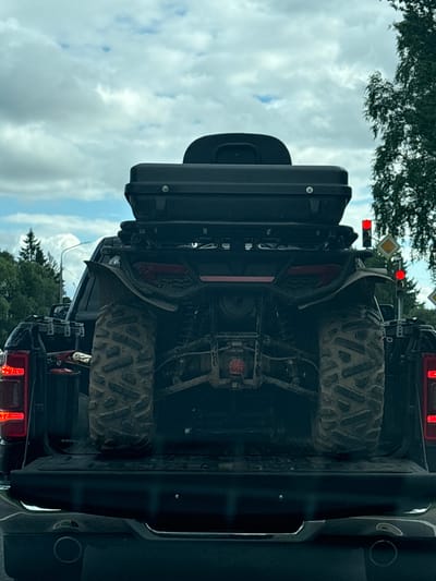 The image shows the rear view of a black pickup truck with an ATV loaded in the truck bed. The ATV is red and black, with large knobby tires covered in mud. A black storage box is mounted on top of the ATV. The truck has red taillights. In the background, there are trees and a cloudy sky. Traffic lights and road signs are visible on the right side of the image. The scene appears to be on a road in a rural or suburban area. The image is taken from inside another vehicle, suggesting the pickup truck is in motion or stopped in traffic. The location is identified as Мазалаўскі сельскі Савет, Belarus.