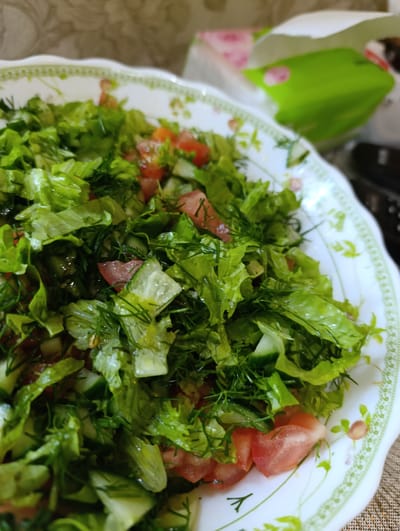 A close-up, slightly high-angle shot captures a serving of fresh salad in a white, oval bowl with a decorative green and floral rim. The salad consists of chopped lettuce, diced tomatoes, and sliced cucumbers, interspersed with finely chopped dill. The ingredients appear moist, suggesting they may have been dressed. In the background, slightly out of focus, is a partially opened white paper bag with a green interior and pink floral patterns. The bag is positioned to the right of the bowl, and beyond it, the environment appears to be an indoor setting with a textured, possibly fabric, surface underneath the bowl. The location of Tugulym, Russia, is not visually evident in the provided image.