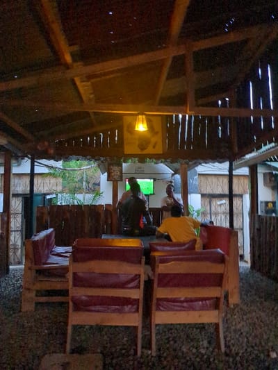 The image shows a dimly lit, outdoor eating area with a wooden roof and a wooden slatted wall in Morogoro, Tanzania (coordinates: -6.835, 37.653). There are four men sitting at wooden tables and chairs. One man is wearing a black shirt and dark pants, another is wearing a white shirt and beige pants, the third man is wearing a yellow shirt and blue jeans and the fourth man is facing away from the camera and is wearing a dark shirt. The tables have red cushions and a television mounted on the wall. A hanging light fixture is visible in the upper right corner of the image. The floor is made of small stones. There are plants in the background.