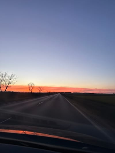 The scene depicts a two-lane asphalt road at sunset or sunrise in Vaglen, Bulgaria. The sky occupies the upper two-thirds of the image, displaying a gradient from a deep lavender-blue at the zenith, transitioning horizontally to intense orange and red hues with defined striations near the horizon. The road extends centrally into the distance, featuring white dashed lane markings and a solid white line on the right shoulder. The foreground includes a dark reflection on what appears to be a vehicle's dashboard or hood, mirroring the sky's orange and red tones. To the left of the road, several leafless deciduous trees are silhouetted against the brightly colored sky, standing on a dark, indistinct verge. To the right, a dark agricultural field stretches towards the horizon. In the far distance on the road, faint points of light indicate the presence of other vehicles. No individuals or specific interactions are discernible. The environment is rural, characterized by open fields and sparse trees under a clear, transitioning sky.