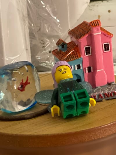 The image shows a close-up, overhead view of a collection of small decorative items on a wooden surface. A Lego minifigure with a yellow head, purple hat, and green torso is lying on its back, with its legs pointing upwards and outwards. The minifigure's face is contorted in what appears to be an expression of surprise or pain, with one eye squinted shut and the mouth open.
To the left of the minifigure is a clear glass orb containing a red, abstract shape that resembles a stylized fish. The orb has small bubbles trapped within it.
Behind the minifigure and to its right is a miniature pink and blue building with a red tiled roof. The building is detailed with windows and a chimney. In front of the building, on the base, the word "RANO" is visible in red lettering.
The wooden surface appears to be a tabletop or shelf. The background is blurred, but suggests a domestic or display setting with some white objects and possibly plastic sheeting. The lighting is somewhat dim, creating shadows that add depth to the scene. There are no discernible features that would place this scene specifically in Maidstone, United Kingdom.