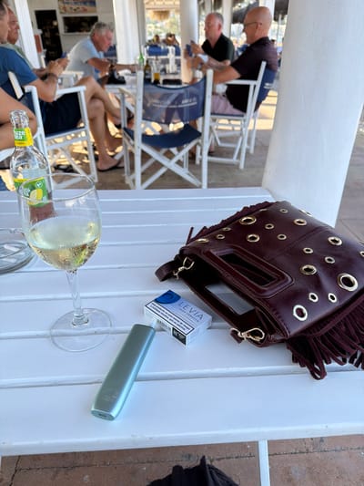 This media file captures a casual outdoor scene, likely at a restaurant or cafe in Xàbia, Spain. The image is oriented sideways. In the foreground, a glass of white wine sits on a white wooden table, next to a partially empty bottle of "fuze tea Limón." A light blue electronic smoking device and a pack of "LEVIA ZERO TOBACCO" cigarettes are also on the table. The text on the cigarette pack states, "Este producto no está exento de riesgo y contiene nicotina, que es adictiva."
Further back, a burgundy handbag with gold eyelets and fringe rests on the table. Several people are visible in the background, seated at tables and chairs, some appearing to be engaged in conversation. One person is shown bending down. The overall impression is of a relaxed daytime gathering. The bright lighting suggests it is a sunny day. The white structure in the background could be part of a building or a patio cover.