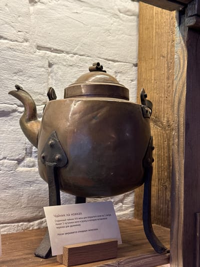 The image shows an antique teapot with a spout, legs, and a handle resting on a wooden ledge. A label next to the teapot reads in Russian about the teapot being from the 19th century, used for open fire, with a capacity of 2 liters. The text also describes the cast iron legs and handle, with a wooden extension that can be inserted. The background includes a rough white brick wall and wooden beams. It appears to be located in the city of Novo-Nikol'skoe, Russia.