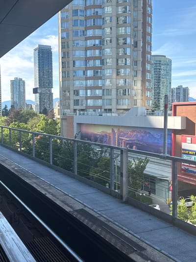 The image captures a view from an elevated walkway, likely a SkyTrain platform, looking towards a cityscape with residential and commercial buildings. The scene is set in Burnaby, Canada, as indicated by the context of SkyTrain stations.

In the foreground, a metallic walkway with a diamond plate surface runs diagonally from bottom left to upper right. A metal railing with vertical posts and horizontal bars separates the walkway from the edge. Below the walkway, train tracks are visible.

Beyond the railing, a dense canopy of green trees stretches across the mid-ground, interspersed with some trees showing fall colors. Behind the trees, a large building features a prominent mural depicting a stylized cityscape with mountains in the background. The mural uses a palette of blues, purples, and oranges.

Further back, several high-rise buildings dominate the skyline. One is a slender, dark glass skyscraper with a distinctive cantilevered section. To its right, a rounded glass tower reflects the sky. Other residential buildings, characterized by numerous windows and light-colored facades, are also visible.

At the street level, a commercial building with a red facade is identifiable, with signage for "BEST BUY" and "Revall." A dark-colored vehicle is parked on the street, and a person is walking near a pedestrian crossing. The sky is clear blue with a few wispy clouds. The overall impression is a bright, sunny day in an urban environment with a mix of modern architecture and natural elements.