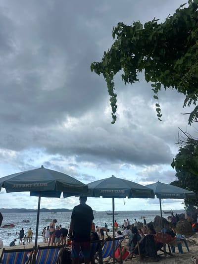 This media file captures a scene on a beach, likely in Phru Nai, Thailand, under an overcast sky. The image is oriented vertically. In the foreground, several blue beach umbrellas with the text "JETSKI CLUB" are visible. These umbrellas provide shade to people sitting on beach chairs, some of whom are partially visible. The people appear to be enjoying a day at the beach, with some engaged in activities near the water.
The middle ground shows the ocean and a shoreline crowded with people. Many are standing or walking along the water's edge, while others are wading or swimming. Beach chairs and umbrellas are scattered along the sand. A few small boats are present in the water. The overall atmosphere suggests a moderately busy beach day, possibly in the late afternoon given the cloudy conditions and diffused light. The presence of the "JETSKI CLUB" umbrellas and the jet skis in the water indicates a recreational area focused on water sports.
The background features a distant coastline with hills or mountains. The sky is filled with heavy, grey clouds, suggesting the possibility of rain or a storm approaching. Despite the clouds, there are glimpses of lighter sky, indicating it is not completely dark. The foliage from trees hangs into the frame from the top left, adding a natural element to the beach scene. The overall mood is relaxed, though the cloudy weather might temper the usual vibrancy of a sunny beach day.