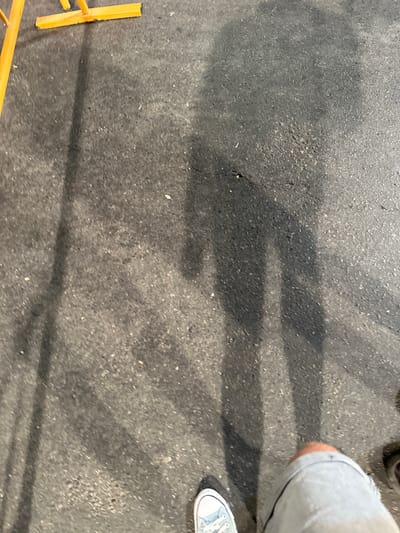 A first-person perspective shot shows a pair of light blue Converse sneakers on asphalt. The subject's legs, clad in light gray shorts, are visible. The ground is a dark, rough asphalt surface with scattered light-colored gravel. Several dark shadows stretch across the asphalt, indicating the presence of people or objects casting them. In the upper left corner, a section of a bright yellow metal structure, possibly scaffolding or a temporary barrier, is visible. The overall impression is an outdoor setting, possibly a public space or construction area. The image does not provide specific visual cues to pinpoint a location within Alicante/Alacant.