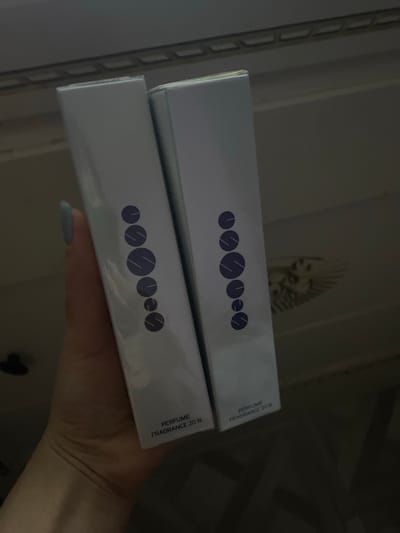 This image, taken in Verkhnevesyoloye, Russia, shows a person's hand holding two identical rectangular boxes, likely packaging for perfume. The boxes are positioned vertically, with one slightly behind and above the other, displaying their side panels to the viewer.

The scene is indoors and dimly lit, suggesting a room environment. In the blurry background, a light-colored wall or surface is visible, featuring an indistinct decorative element that appears to be a stylized head or figure, possibly resembling a sphinx or a creature with a prominent headdress. A vertical structure, potentially a shelf or part of furniture, is also discernible on the left side of the background.

The hand holding the boxes has neatly manicured fingernails painted with a light, pastel blue or grey polish. Both boxes are white or light grey and appear to be made of cardboard, sealed as if they are new products. Each box features a distinct logo on its front panel, composed of six dark blue or purple circular shapes, each bisected by two diagonal lines, arranged horizontally. Vertically printed on the right side of the visible panels of both boxes is the text: "PERFUME FRAGRANCE 20%". The activity depicted is simply the presentation or holding of these two product boxes. No specific emotions are discernible.