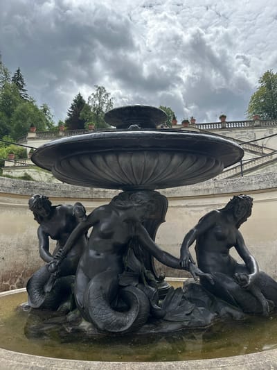 The image captures a bronze fountain sculpture depicting three mermaids in a pool of water. The mermaids are arranged closely together, with their tails intertwined. The sculpture is set against a backdrop of a light-colored, textured wall and a curved stone staircase with potted plants along its upper railing. Beyond the staircase, there are trees and a dramatic cloudy sky. The fountain is located in Ettal, Germany, likely within the grounds of Ettal Abbey. The lighting suggests an overcast day.