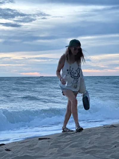 This media file captures a person on a beach in Tong Yang, Thailand. The subject, a woman, is seen walking near the water's edge, with waves crashing on the shore. She is wearing a white tank top, shorts, a green cap, and sandals. She is also carrying a bag over her shoulder and holding a clear bottle. The setting is a beach, with sand in the foreground and the ocean stretching out to the horizon. The sky is visible, with clouds suggesting it might be late afternoon or early evening, given the soft light and pastel colors on the horizon. The overall mood appears relaxed and casual, indicative of a beach outing. No visible text is present in the image.