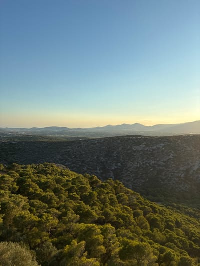A panoramic landscape view captures the rolling hills and mountains of Limenas Markopoulou, Greece, bathed in the warm glow of late afternoon sun. The sky is a clear, gradient blue, transitioning to a soft yellow near the horizon. In the foreground, a dense forest of green pine trees covers a steep slope, their tops illuminated by sunlight. Beyond the pine-covered hills, a rugged, rocky terrain is visible, also sparsely vegetated. Further in the distance, a hazy expanse of mountains stretches across the horizon, with hints of a distant town or settlement visible between them. The overall scene evokes a sense of natural tranquility and the vastness of the Greek landscape.