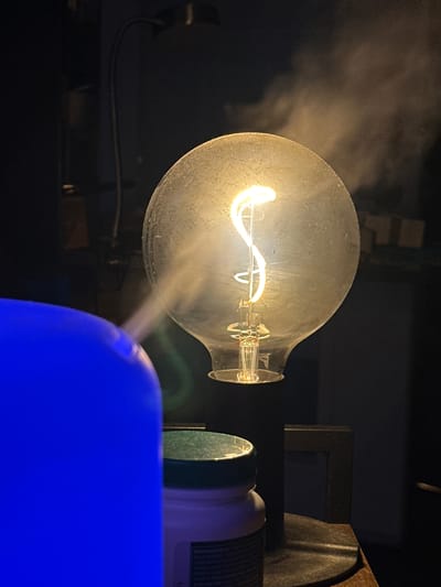 The image captures a close-up of an illuminated incandescent light bulb with a visible filament, emitting a warm, yellow glow. A stream of vapor or mist is seen rising from the right side and dissipating into the dark background. In the foreground, a large, bright blue object, possibly a diffuser or humidifier, dominates the left side of the frame. Partially visible behind it is a white container with a green lid, which appears to be a jar or bottle. The scene is dimly lit, with the light bulb and the blue object serving as primary light sources. The background is dark and indistinct, suggesting an indoor setting, likely a dimly lit room. There are no visible people or animals. The composition is tightly framed, focusing on the interplay of light, mist, and the foreground objects. The location in McMasterville, Canada, is not discernible from the visual elements present in the image.