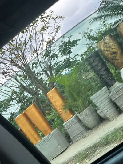 The image displays an outdoor scene in Elimgbu, Nigeria, viewed through a vehicle's side window, evidenced by the dark frame in the foreground and reflections on the glass. A row of large planters and decorative elements is arranged on a light-colored ground surface, likely dirt or weathered concrete, extending diagonally from the lower left to the upper right. These objects include tall, rectangular structures with a shimmering gold finish, one featuring distinct circular indentations. Adjacent to these are multiple gray concrete planters with varying textures, such as ribbed patterns and embossed designs. Several planters contain live green plants, consistent with shrubs or small trees. In the background, dense tropical vegetation, including a large tree with sparse leaves and numerous green-leafed trees, stands beneath an overcast or heavily clouded sky. No individuals or discernible activities are present.