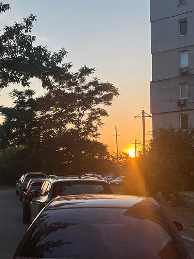 This is a vertical photograph taken at an eye-level perspective, capturing a sunset scene in Kropyvnytskyi, Ukraine. The sun is setting, casting a warm orange glow across the sky. In the foreground, the side of a dark blue car is visible, reflecting the colors of the sunset and the surrounding trees. Behind the car, a row of parked vehicles can be seen.
Further in the background, silhouetted against the bright sky, are trees and utility poles with electrical wires. An apartment building, characterized by its grey facade and visible windows, is present on the left side of the image, with some units appearing to have air conditioning units installed. The overall scene suggests a residential area at dusk. The lighting indicates it is late afternoon or early evening, and the sky is clear, suggesting fair weather. There is no visible text in the image.