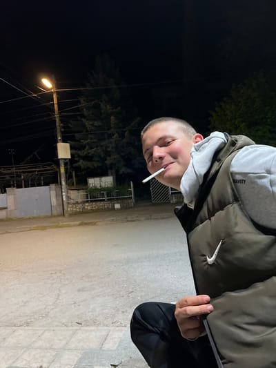 A young man stands on a paved surface in what appears to be a street or sidewalk. He is wearing a grey hoodie under a dark green puffer vest and black pants. He has a very short haircut and is holding a cigarette in his mouth.
The background is dark, with a street light illuminating a tree and some buildings. A utility pole with lines is also visible. Based on the information provided, the location is Karavelovo, Bulgaria.